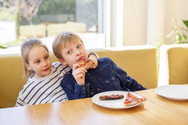 Güneşli kapalı masada pizzayı paylaşan iki çocuk, bir çocuk şakacı bir şekilde diğerine yaslanıyor..