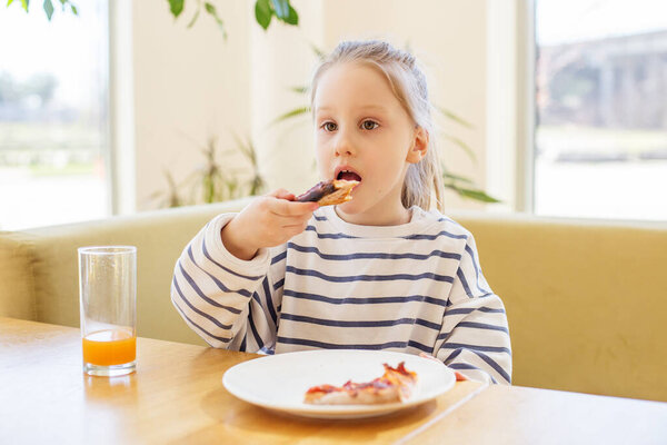 Kid eating a slice of pizza while sitting at a sunny indoor table with glass of orange juice.