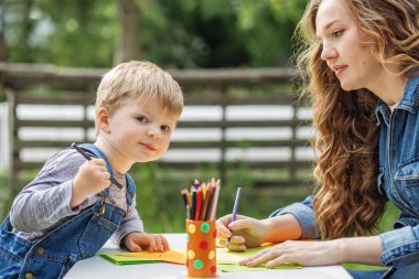 Anne ve küçük oğlu güneşli bir günde açık hava masasında boyama faaliyetlerinin keyfini çıkarıyorlar..