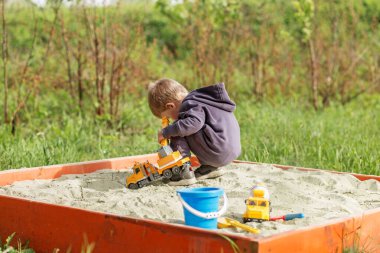 Kapüşonlu çocuk kum havuzunda oyuncak kamyonlarla oynuyor, açık hava parkında güneşli bir günün tadını çıkarıyor..