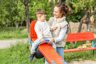 Anne ve çocuğun parkta renkli tahterevallide gülüp oynadığı neşeli bir an..