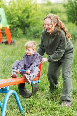 Yeşil eşofmanlı anne ve genç oğlu, yemyeşil parkta renkli tahterevallide neşeli anların tadını çıkarıyorlar..