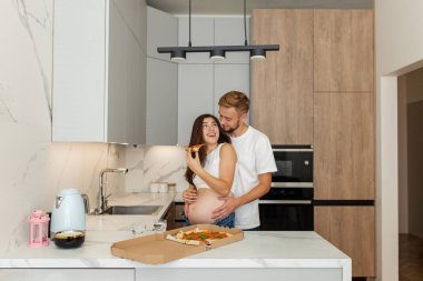 Sevgi dolu çift modern mutfakta hassas bir anı paylaşıyor. Hamile kadın pizzanın tadını çıkarırken eşi onu kucaklıyor..