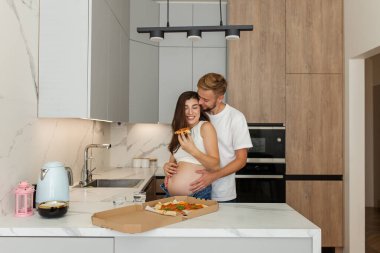 Hamile kadın ve partneri modern mutfakta samimi bir anı paylaşıyor. Pizzanın tadını çıkarırken sıcak bir şekilde sarılıyorlar..