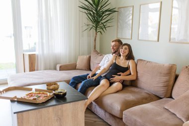 Neşeli hamile kadın ve partneri güzel ışıklandırılmış oturma odasındaki büyük bölmeli kanepede dinleniyor, pizza ve atıştırmalıklarla keyifli bir hafta sonu geçiriyorlar..