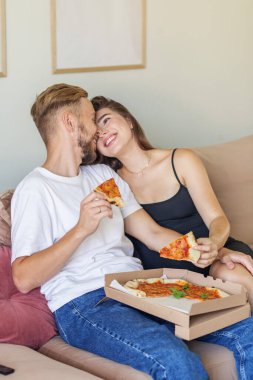 Neşeli çift kanepeye yakın otururken pizzayı paylaşmayı sever. Evdeki aşk ve mutluluk anlarını yakalar..