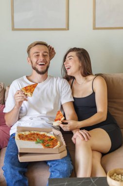 Mutlu çift gülüyor ve pizzanın tadını çıkarırken rahat koltukta oturup, evde mutlu ve rahat bir anı paylaşıyorlar..