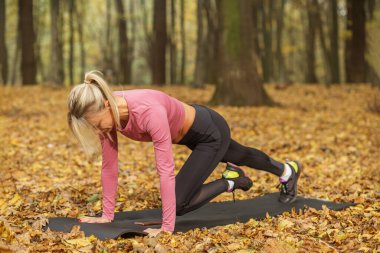 Pembe bluzlu ve siyah taytlı bir kadın ormanda yoga minderiyle dağcılık yapıyor. Açık hava antremanı sırasında içi sonbahar yapraklarıyla dolu..
