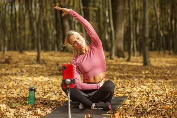 stock image Fit woman in pink top and black leggings demonstrating side stretch during yoga routine while filming with smartphone in an autumn forest.