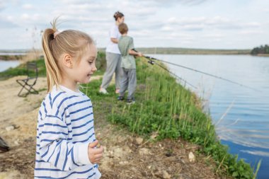 Genç kız nehir kıyısında duruyor, ailesinin sakin, güneşli bir günde arka planda balık tutması gibi gülümsüyor..