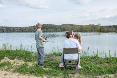 Anne kızını öperken, oğlu da gölün huzurlu kıyısında balık tutuyor. Dışarıda sakin bir günün tadını çıkarıyor..