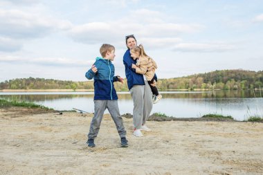 Anne, huzurlu göl kenarında iki çocukla neşeli bir anı paylaşıyor, birlikte gülüyor ve dışarıda eğleniyor..