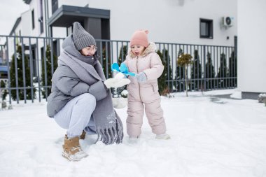 A mother kneels in fresh snow, smiling at toddler who holds a playful snowball ready to throw. Their cozy winter outfits contrast with the white landscape. clipart