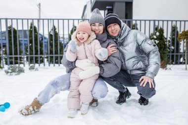 Karda mutlu bir aile oynuyor, kalın giysiler içinde. Ebeveynler kameraya neşe içinde poz veren genç kızlarının yanında diz çöker. Evleri rahat bir zemin ekliyor..