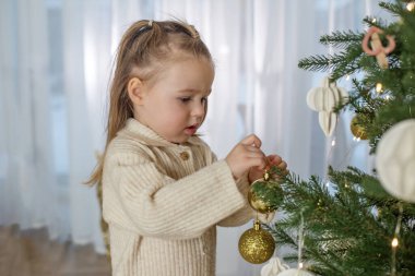 Çocuk, yumuşak ışık ve bayram neşesiyle dolu sıcak, kapalı bir ortamda, güzel dekore edilmiş Noel ağacına altın süsler takıyor..