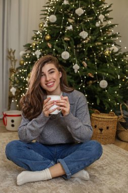 Genç bir kadın bağdaş kurup yerde oturuyor, elinde sıcak bir içecek var. Arkasındaki şenlikli Noel ağacı ışık ve süslerle süslenerek rahat bir atmosfer yaratıyor..