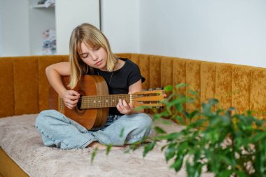 Çocuk konforlu kanepeye bağdaş kurarak oturur, ışık dolu neşeli yaşam alanında gitarını tıngırdatmaya odaklanır..