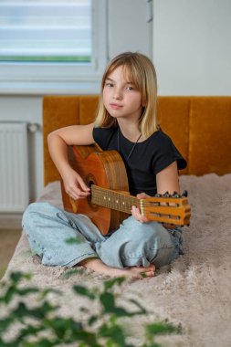 Tween rahat bir yatakta bacak bacak üstüne atarak oturur, evde huzurlu bir öğleden sonra geçirirken gitar tıngırdatır..