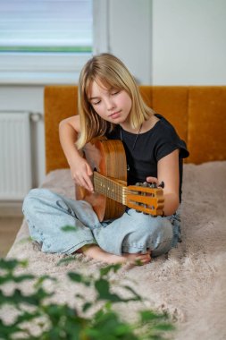 Tween yatakta bacak bacak üstüne atmış, parlak ve davetkar odada ustalıkla gitar çalıyor. Yumuşak ışık pencerelerden süzülür.