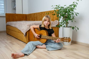 Genç bir kız yerde bağdaş kurup gitarını parlak, davetkar bir mekanda tıngırdatıyor. Rahat bir kanepe ve bitki sıcaklık ekler..