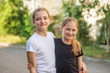 İki çocuk, yemyeşil bir mahallede açık mavi gökyüzünün altında parlak bir şekilde gülümseyerek birbirlerine yakın duruyorlar. Mutlu ve kaygısız görünüyorlar, sıcak bir günde birbirlerinin arkadaşlığından zevk alıyorlar..