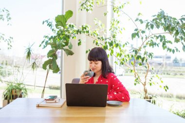 Kırmızı bluzlu bir kadın laptopuna odaklanırken sıcak bir içecek yudumlarken gülümsüyor. Güneş ışığı, yeşilliklerle çevrili alanı doldurur ve üretkenlik için rahat bir atmosfer yaratır..