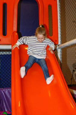 Neşeli gülüşü olan küçük bir çocuk, kapalı bir oyun alanında canlı turuncu kaydırak kayarak aşağı kayıyor. Parlak renkler ve yumuşak yüzeyler çocuklar için eğlenceli bir ortam yaratır.
