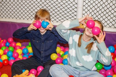 Two children are enjoying their time in a vibrant ball pit, playfully holding colorful balls up to their eyes. They laugh and spread joy in a lively indoor playground setting. clipart