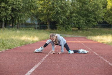 A tween in gray clothing and protective gear has fallen while roller skating on a red outdoor track. She appears determined as she stretches forward, surrounded by greenery in the background. clipart