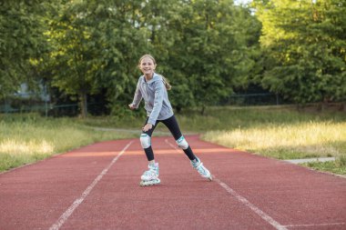 Genç bir kız, parkta pürüzsüz kırmızı bir pistte kayarken, süzülerek gülümsemekten zevk alır. Güneş parlıyor yemyeşil yemyeşil parlıyor.
