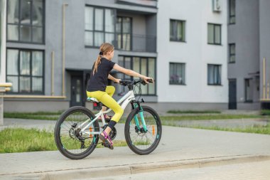 At kuyruklu bir kız şehir merkezinde kaldırım boyunca bisikletini sürüyor. O, öğleden sonra temiz havanın tadını çıkarırken siyah bir tişört ve parlak sarı taytlar giyer..