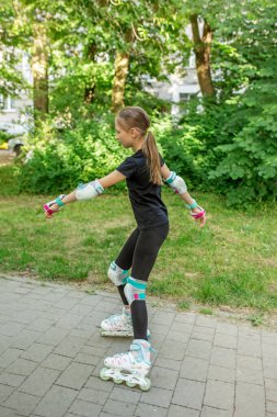 Genç bir kız parkta paten kayıyor. Denge ve tekniğine odaklanırken koruyucu giysi giyer. Ilık havanın tadını çıkarırken çevresini çevreleyen canlı yeşillik.