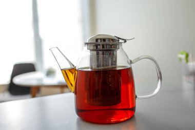 Kehribar çayıyla dolu berrak bir cam çaydanlık beyaz bir tezgahta durur. Güneş ışığı çaydanlığı aydınlatır, çayın zengin rengini ve içindeki ağ zerk ediciyi vurgular..