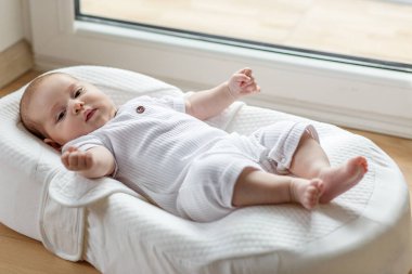Beyaz elbiseli tombul bir bebek, yastıklı bir bebek odasında, pencerenin yanında, rahat ve mutlu görünüyor.