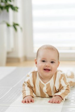 Çizgili kıyafetli gülümseyen bebek evde yumuşak bir oyun minderinde göbek yapıyor, doğal ışıkta motor becerilerini geliştiriyor..