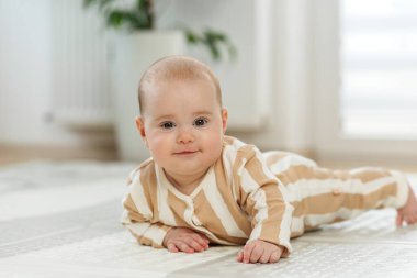 Çizgili kıyafetli gülümseyen bebek evde yumuşak bir oyun minderinde göbek yapıyor, doğal ışıkta motor becerilerini geliştiriyor..