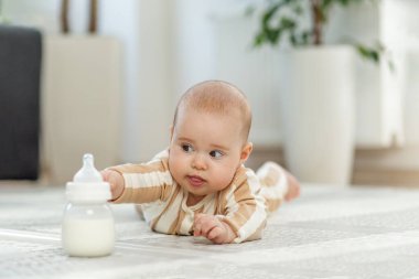 Çizgili giysiler içinde karnının üzerine uzanmış meraklı bir bebek, parlak bir odada yumuşak bir oyun paspasının üzerindeki süt şişesine uzanıyor..