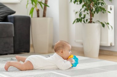 Meraklı bir bebek yumuşak bir halıya uzanır, çağdaş oturma odasına bakarken renkli bir diş çıkarma oyuncağı tutar. Yeşil bitkiler ve minimalist dekor rahat atmosfere eklenir.