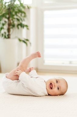 Neşeli bir bebek yumuşak bir yüzeyde uzanıyor, ayaklarını tutuyor ve gülümsüyor. Güneş ışığı geniş pencerelerden süzülür, kapalı mekanın neşeli atmosferini aydınlatır..