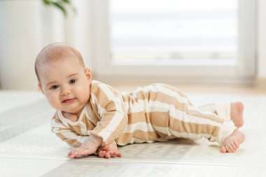 Çizgili pijamalarıyla gülümseyen bebek, parlak bir evde minderli bir oyun paspası üzerinde yuvarlanmayı öğreniyor..
