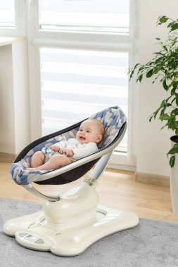 Neşeli bir bebek güneşli bir odada modern bir salıncakta dinleniyor. Yumuşak renkler ve doğal ışık sakin bir atmosfer yaratırken, yumuşak hareket rahatlık sağlar..
