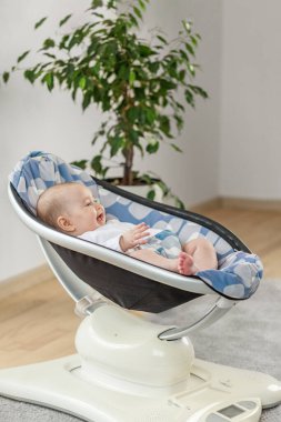 Neşeli bebek evde yastıklı, modern elektrikli bebek rockçısında yatıyor, saksı bitkisinin yanında hareketin ve konforun keyfini çıkarıyor..