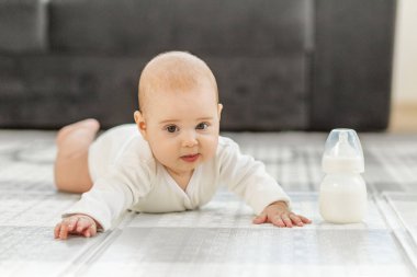 Bebek sıcak bir oturma odasında yumuşak bir halının üzerinde sürünüyor. Çocuk merakla ve heyecanla çevrelerini keşfederken bir süt şişesi yakınlarına yerleştirilir..
