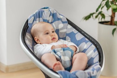 Şirin bir bebek evdeki şık bir koruma koltuğunda rahat bir şekilde oturur. Etrafı aydınlık bir evle çevrilidir. Çocuk meşgul ve rahatlarken mutlu görünüyor..