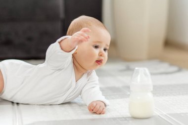 Beyaz giysili meraklı bir bebek yumuşak bir oyun hasırının üzerinde sürünüyor ve yakınlarda bir süt şişesine uzanıyor. Sabah güneşi sıcacık oturma odasını dolduruyor, sıcak bir atmosfer yaratıyor..