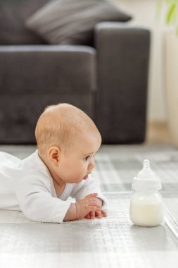 Meraklı bir bebek yerde yatar, yanına konmuş bir şişe süte doğru uzanır. Yumuşak ışık odayı dolduruyor, neşeli keşifler için sıcak ve misafirperver bir atmosfer yaratıyor..