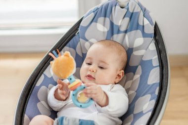 Bir bebek rahat bir fedaide oturmuş renkli bir oyuncağı keşfetmeye odaklanmış. Güneş ışığı odayı dolduruyor, bebeğin tuhaf ifadelerini ve neşeli çevresini vurguluyor..