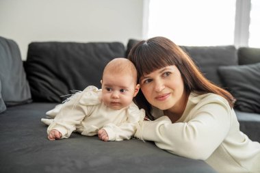 Gülümseyen anne bebeğini karanlık bir kanepede uzanmış, neşeli ve samimi bir ebeveyn anını doğal ışıkta paylaşıyor..