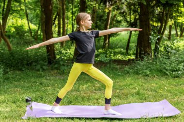 Atletik kıyafetli genç kız yeşil orman ortamında yoga minderinde savaşçı pozu veriyor. Denge ve farkındalığa odaklanıyor..