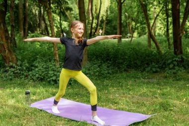 Atletik kıyafetli genç kız yeşil orman ortamında yoga minderinde savaşçı pozu veriyor. Denge ve farkındalığa odaklanıyor..